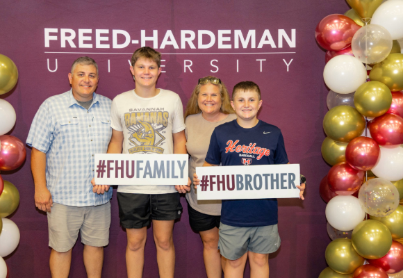 FHU Welcome Home family in front of photo backdrop holding signs with FHU related Hashtags on them