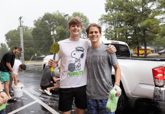 two male student posing with their car cleaning tools in front of a car being washed by 3 other male students