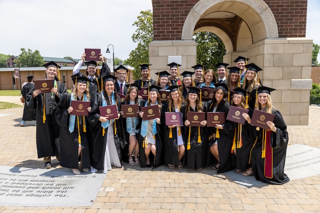 FHU graduates display their diplomas in front of the Bucy bell tower.