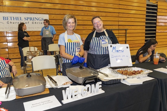 Women wearing aprons serve bacon on a stick at Welcome Home community fair.